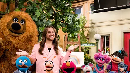 Riccardo Simonetti posiert mit den Figuren aus der "Sesamstraße". Er trägt ein pinkes Outfit und eine goldene Kette.