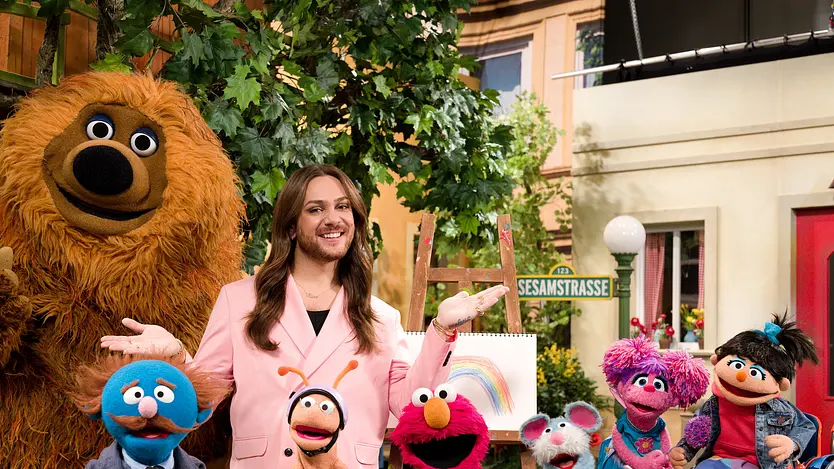Riccardo Simonetti posiert mit den Figuren aus der "Sesamstraße". Er trägt ein pinkes Outfit und eine goldene Kette.