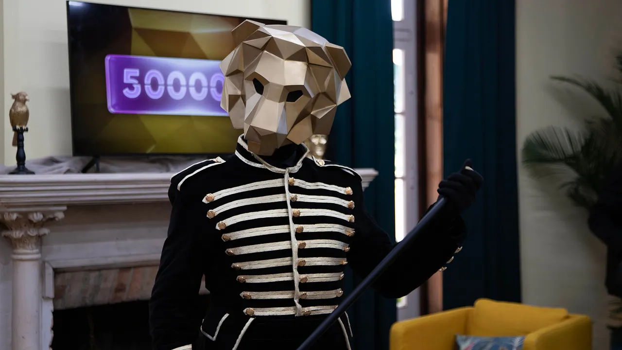 Der Löwe aus "The 50" Eine Person mit goldener Löwenmaske hält einen schwarzen Gehstock und trägt ein golden verziertes, schwarzes Oberteil. Hinter ihr sehen wir einen Fernseher, auf dem in einem lila Kasten die Zahl 500.000 steht. Die Löwenmaske verdeckt die letzte Null. - Foto: Prime Video