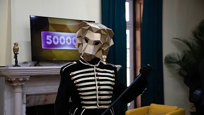 Eine Person mit goldener Löwenmaske hält einen schwarzen Gehstock und trägt ein golden verziertes, schwarzes Oberteil. Hinter ihr sehen wir einen Fernseher, auf dem in einem lila Kasten die Zahl 500.000 steht. Die Löwenmaske verdeckt die letzte Null.  