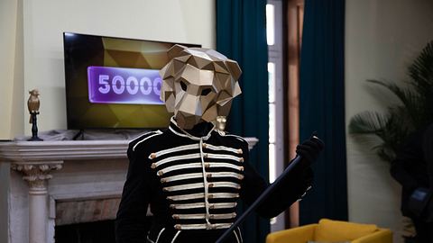 Eine Person mit goldener Löwenmaske hält einen schwarzen Gehstock und trägt ein golden verziertes, schwarzes Oberteil. Hinter ihr sehen wir einen Fernseher, auf dem in einem lila Kasten die Zahl 500.000 steht. Die Löwenmaske verdeckt die letzte Null.   - Foto: Prime Video