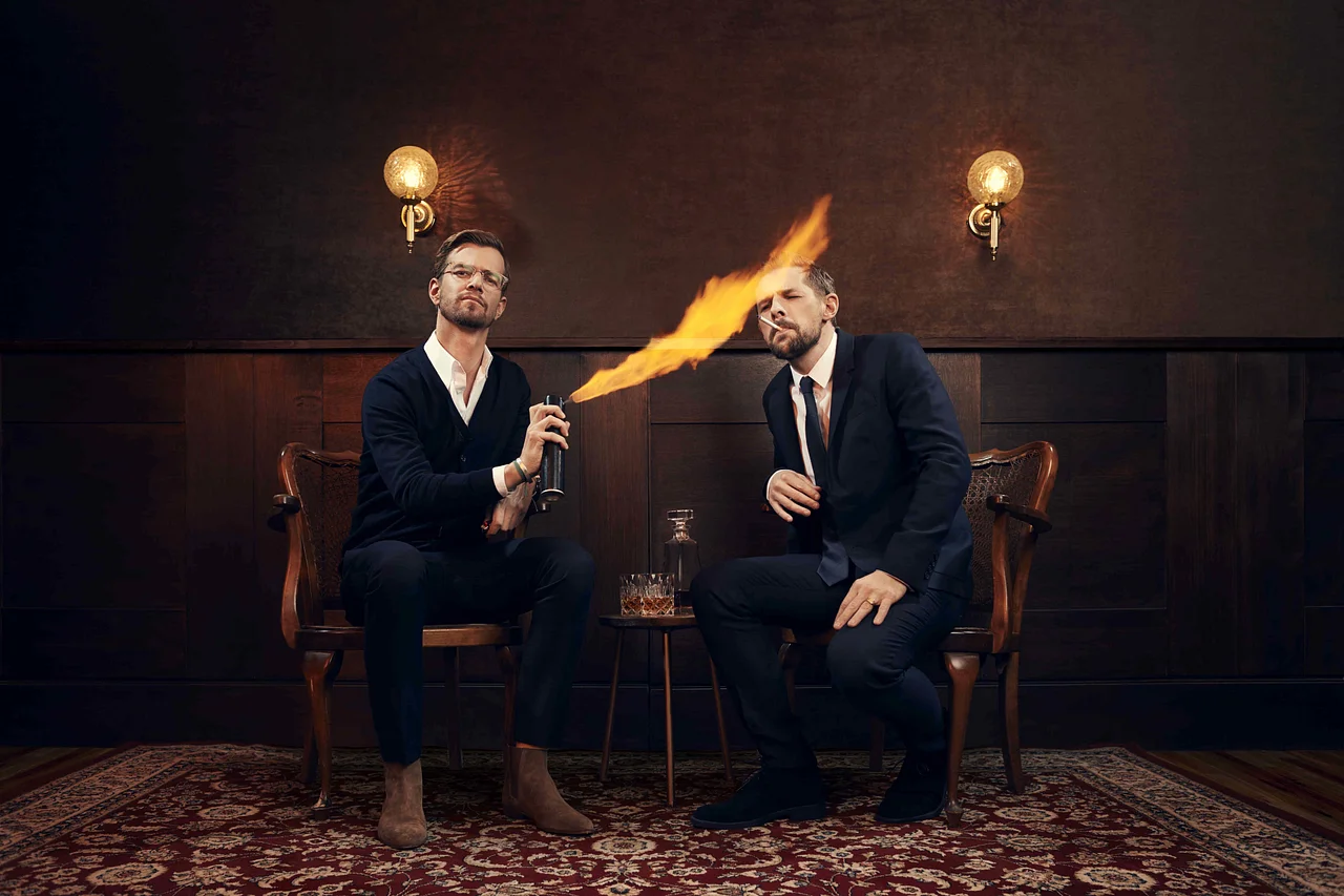 Joko und Klaas sitzen auf Holzstühlen. Joko zündet Klaas Zigarette mit einer brennenden Spraydose an. - Foto: ProSieben