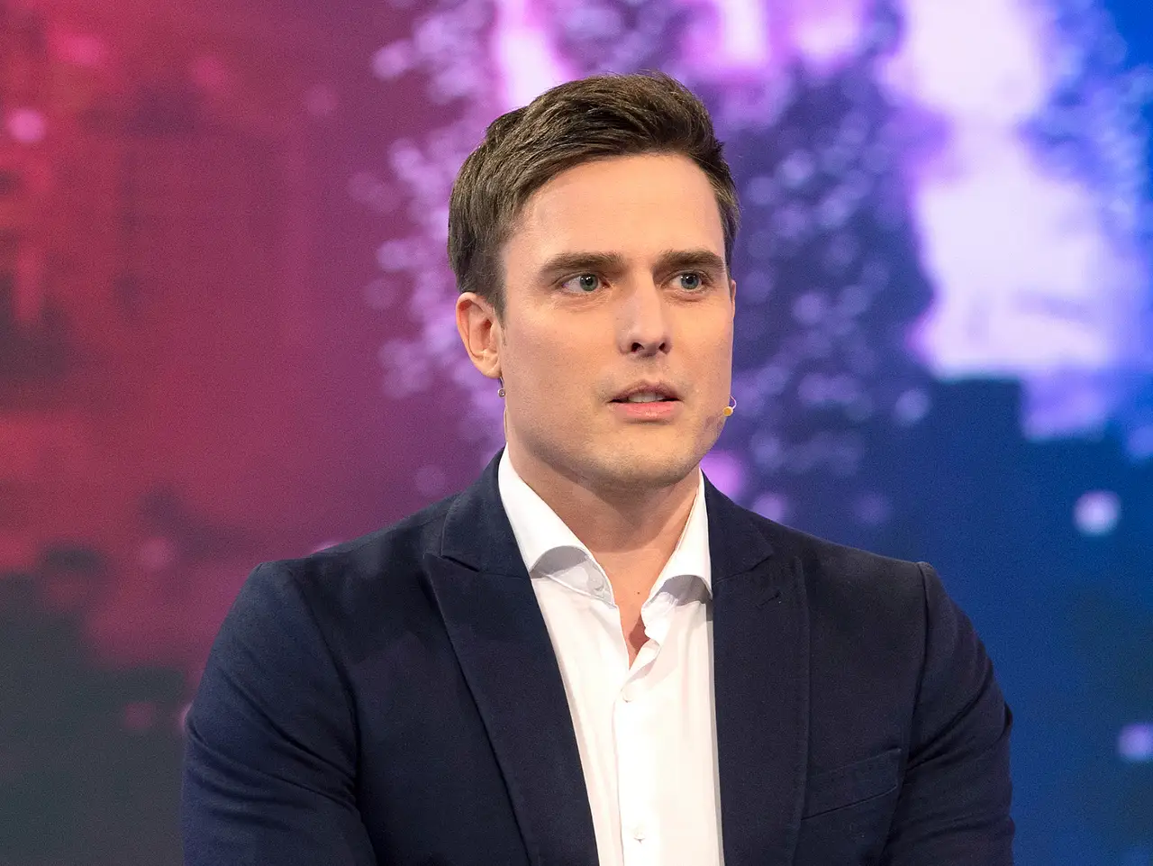 Constantin Schreiber gönnt sich erstmal eine Pause. Constantin Schreiber blickt an der Kamera vorbei. Er trägt einen Anzug, hinter ihm ist eine bunte LED-Wand. - Foto: © WDR/Melanie Grande