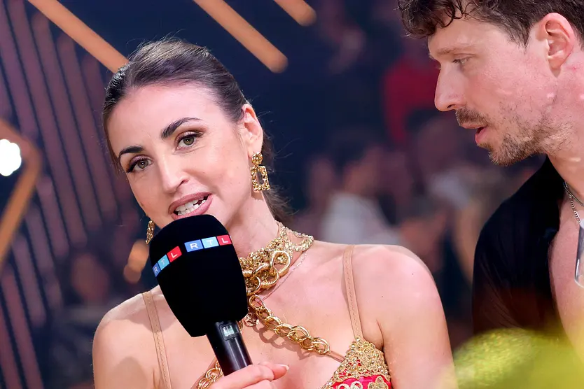Ekaterina Leonova, hier im roten Kleid und dicker goldener Kette, spricht mit angestrengtem Blick in ein RTL-Mikrofon, Valentin Lusin schaut sie erschrocken an. 
