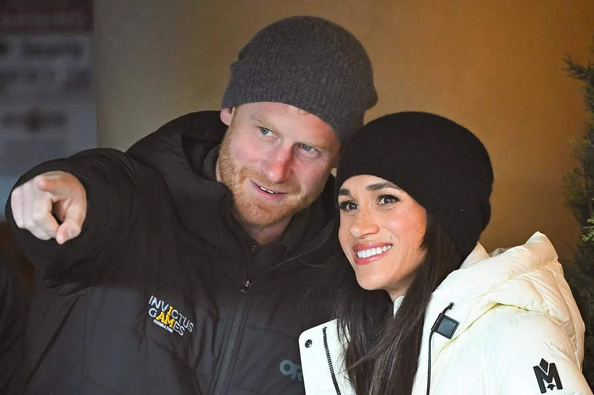 Prinz Harry zeigt mit dem rechten Zeigefinger vor sich, Meghan blickt ebenfalls lächelnd nach vorne.