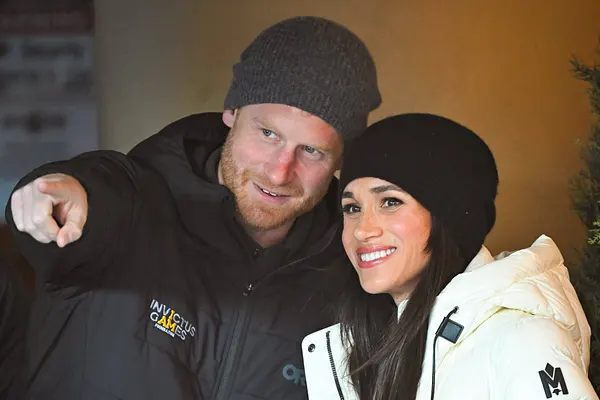 Prinz Harry zeigt mit dem rechten Zeigefinger vor sich, Meghan blickt ebenfalls lächelnd nach vorne.
