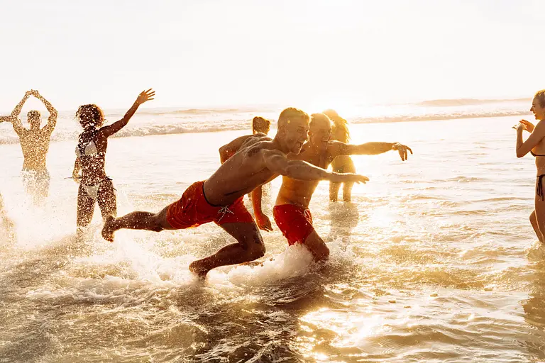 Mehrere Männer und Frauen tummeln sich bei Sonnenuntergang im seichten Wasser am Strand.