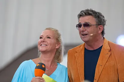 Andrea Kiewel und Hans Sigl stehen nebeneinander und moderieren gemeinsam den "ZDF Fernsehgarten". Kiewel hält ein ZDF-Mikrofon.