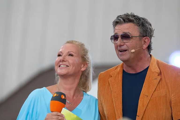 Andrea Kiewel und Hans Sigl stehen nebeneinander und moderieren gemeinsam den "ZDF Fernsehgarten". Kiewel hält ein ZDF-Mikrofon.