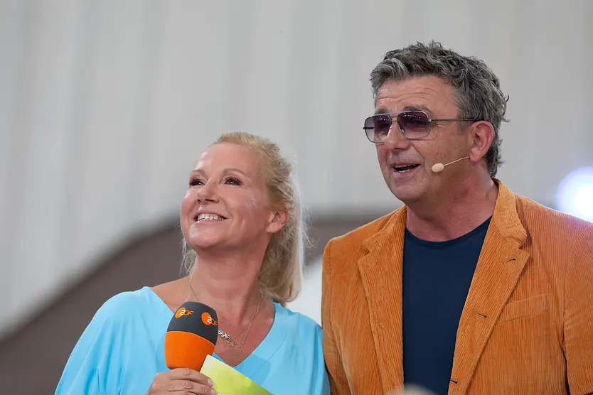 Andrea Kiewel und Hans Sigl stehen nebeneinander und moderieren gemeinsam den "ZDF Fernsehgarten". Kiewel hält ein ZDF-Mikrofon.