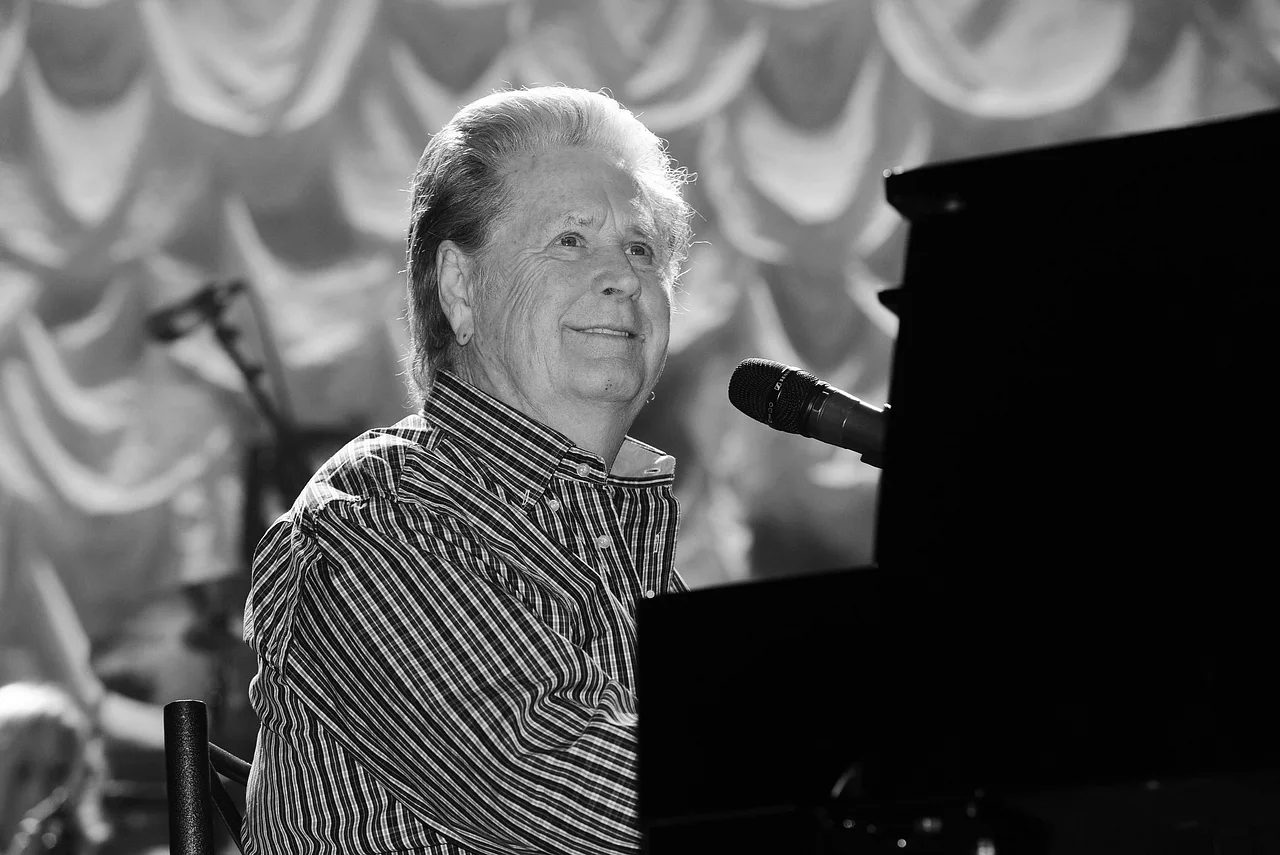 brian-wilson-beach-boys-tot-imago822134473 Schwarzweißfoto von Brian Wilson. Er sitzt lächelnd vor einem Klavier und einem Mikrofon. - Foto: IMAGO / ZUMA Press Wire / Brent Perniac