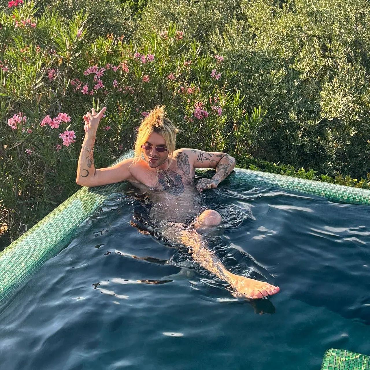 ddp_25876847 Bill Kaulitz sitzt entspannt im Pool, trägt eine Sonnenbrille und zeigt mit der Hand ein Peace-Zeichen. Sein tätowierter Oberkörper ist sichtbar, die blonden Haare sind zum Knoten gebunden. Im Hintergrund blühen rosa Oleandersträucher und grüne Büsche. - Foto: ddp socialmediaservice