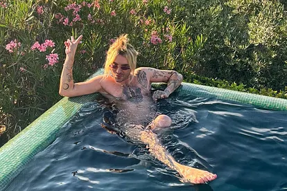 Bill Kaulitz sitzt entspannt im Pool, trägt eine Sonnenbrille und zeigt mit der Hand ein Peace-Zeichen. Sein tätowierter Oberkörper ist sichtbar, die blonden Haare sind zum Knoten gebunden. Im Hintergrund blühen rosa Oleandersträucher und grüne Büsche.
