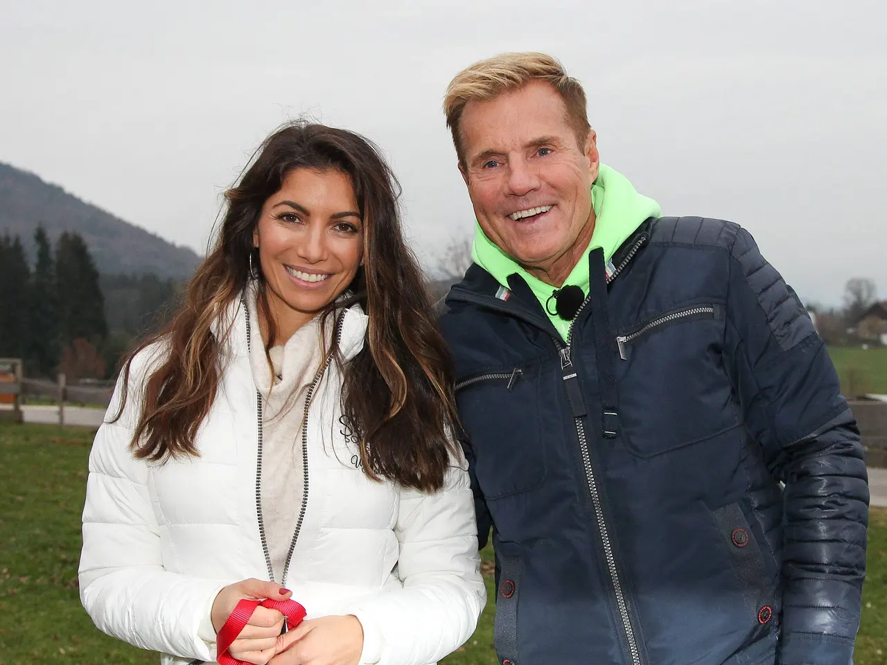 Dieter Bohlen und Carina Walz stehen lächelnd nebeneinander im Freien vor einer Berglandschaft und tragen Winterjacken
