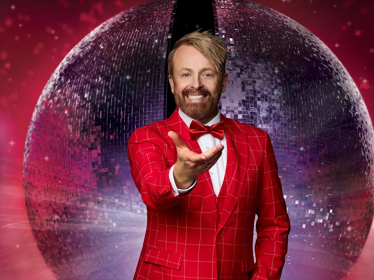 Ross Antony steht vor einer großen glitzernden Discokugel im roten Anzug mit Fliege und streckt lächelnd die Hand aus.