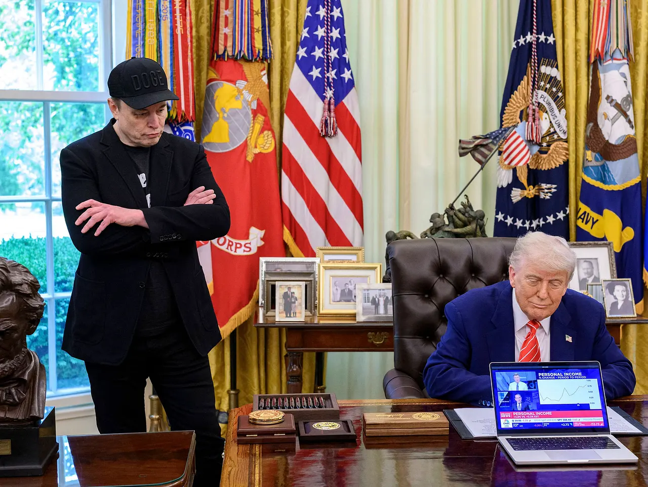 Elon Musk steht mit verschränkten Armen neben dem im Oval Office sitzenden Donald Trump. Heute sind sie keine Freunde mehr. - Foto: IMAGO / ZUMA Press Wire / Molly Riley