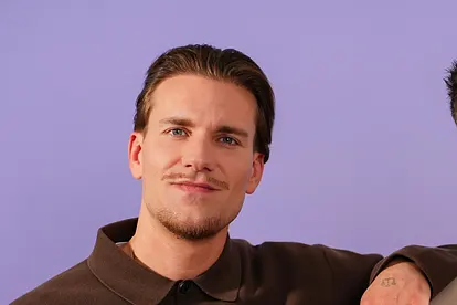 Bachelor Felix Stein lehnt sich vor lilafarbenem Hintergrund lässig auf die Schulter von Martin Braun.