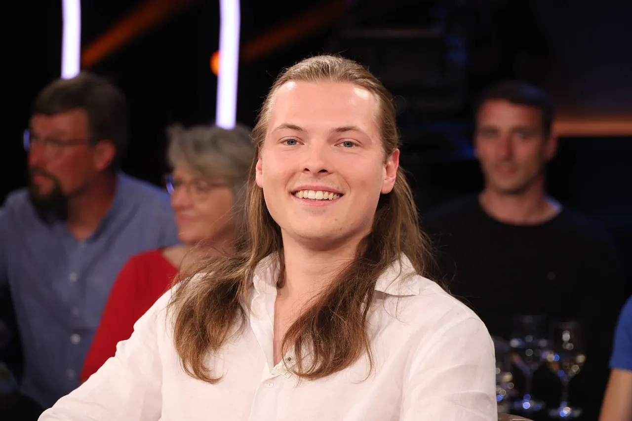 Gabriel Kelly trägt ein weißes Hemd und lächelt in einer Talkshow breit in die Kamera. - Foto: IMAGO / Stephan Wallocha