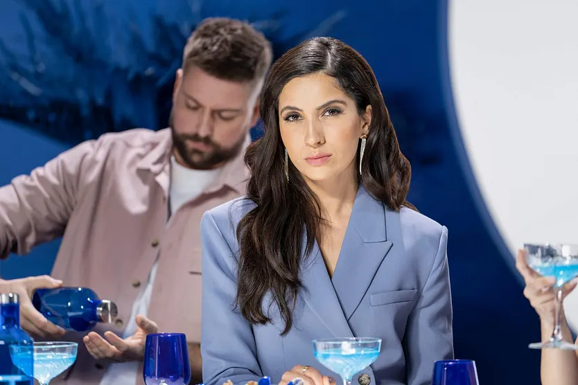 GZSZ-Szene: Laura-Darstellerin Chryssanthi Kavazi sitzt vor einem Drink und guckt ernst, im Hintergrund mixt John-Mime Felix von Jascheroff einen Cocktail.