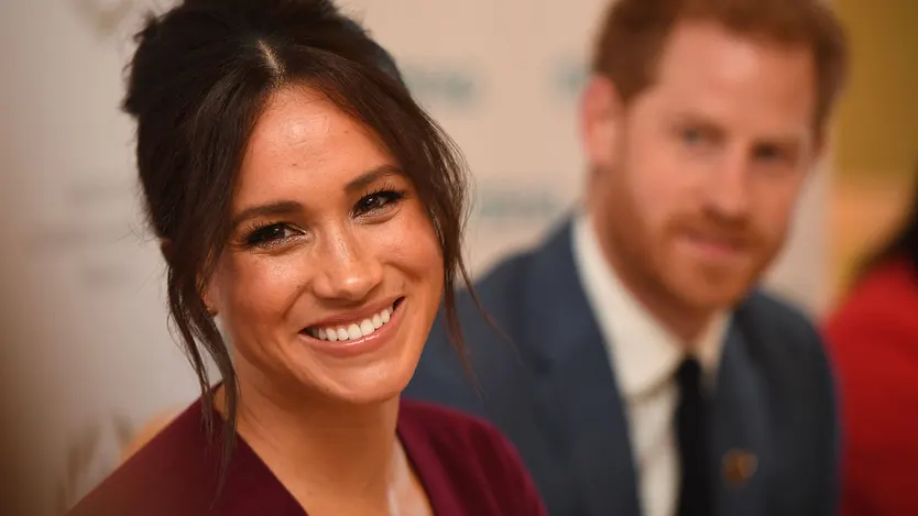 Meghan mit Hochsteckfrisur lächelt. Prinz Harry ist im Hintergrund