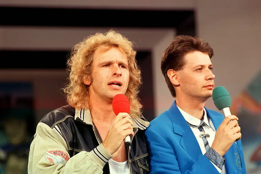 Man sieht Thomas Gottschalk und Günther Jauch Anfang der 90er beim Moderieren. Gottschalk trägt eine Collegejacke, während Jauch einen blauen Anzug aufträgt.
