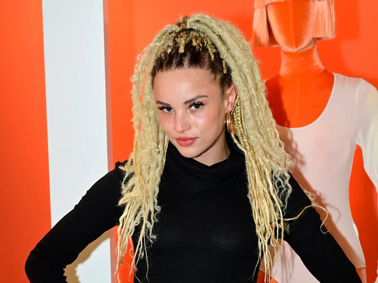 Christin Okpara mit blonden Braids und schwarzem Outfit vor einem rot-weißen Hintergrund, während eines öffentlichen Auftritts. In ihrer Instagram-Story verrät sie jetzt, wieso sie bei Kampf der Realitystars so plötzlich verschwunden ist.  - Foto: IMAGO / pictureteam