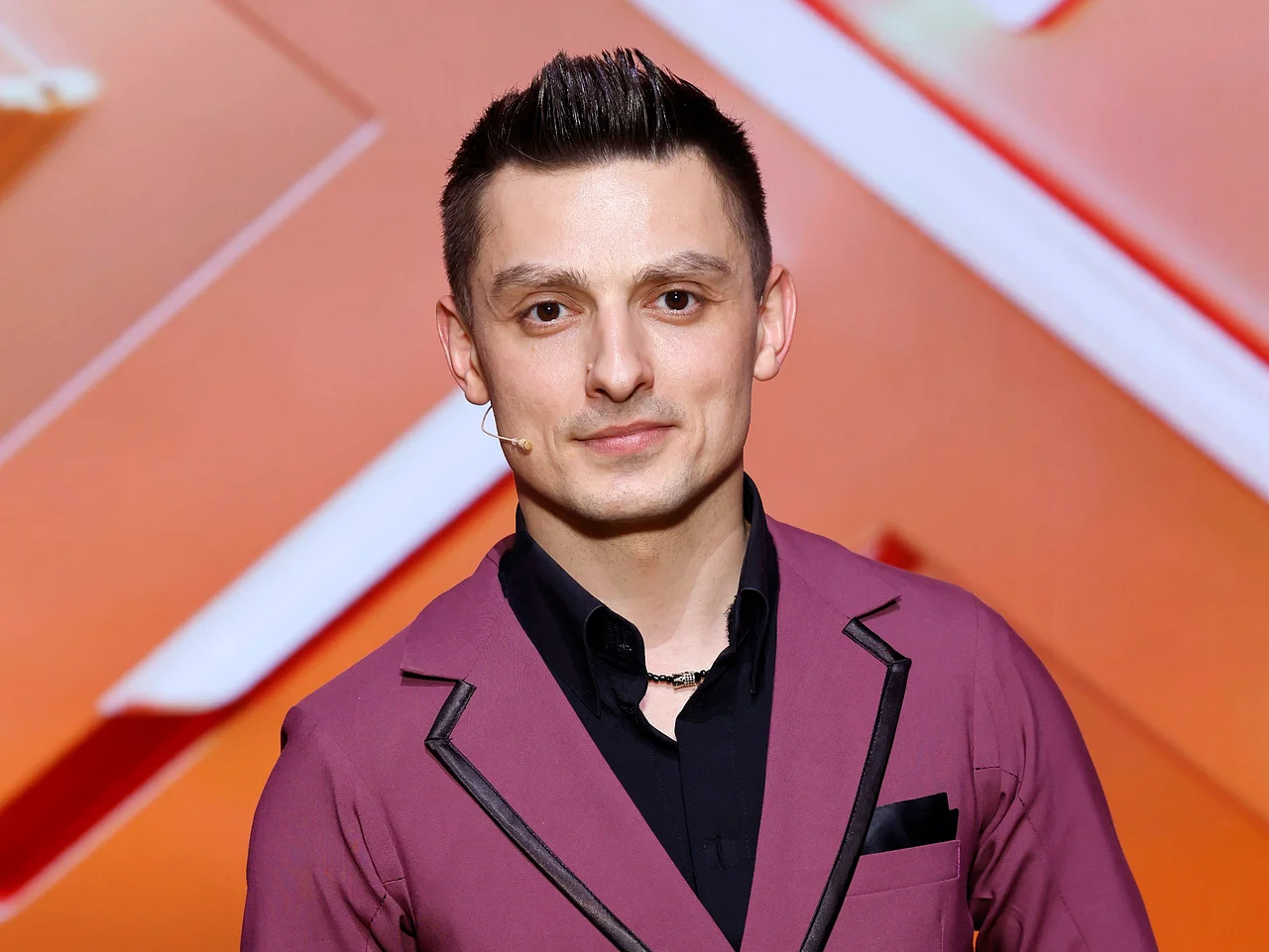 imago806817049 Zsolt Sándor Cseke steht vor einem orangenem Hintergrund und grinst in die Kamera.