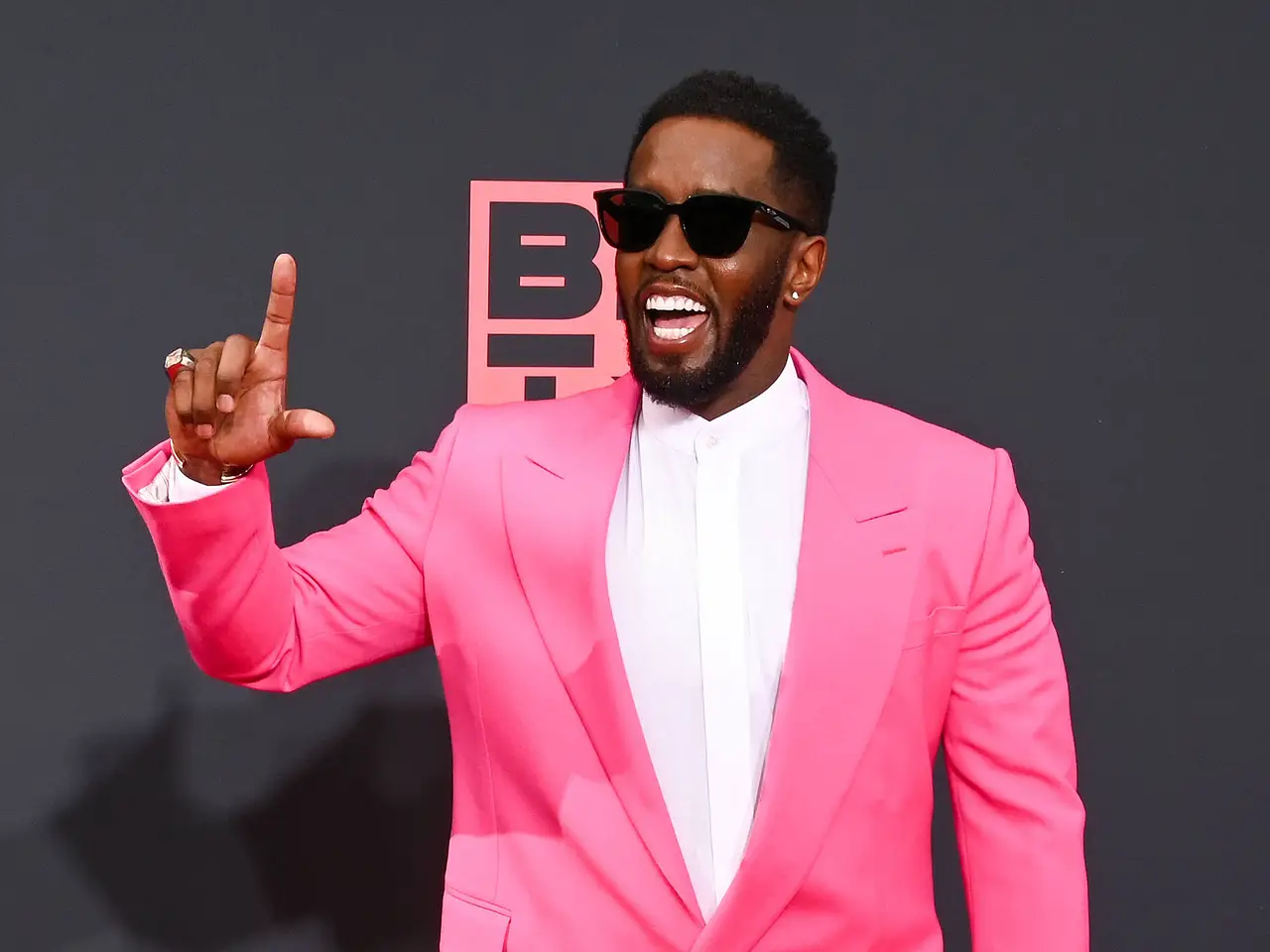 Sean Combs auf dem roten Teppich. Er lacht und trägt eine Sonnenbrille, mit Daumen und Zeigefinger der rechten Hand formt er ein L.