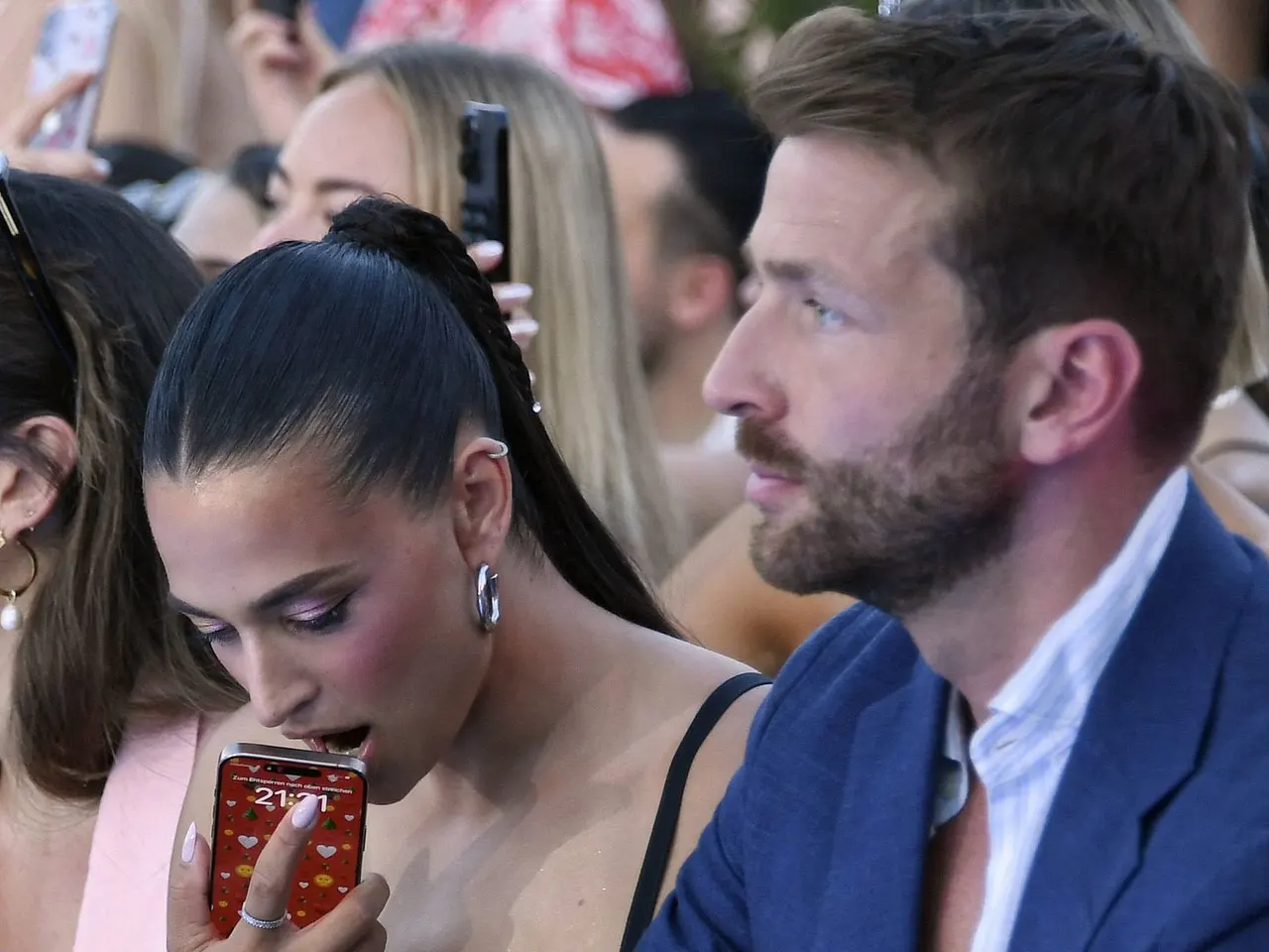 Amira Aly starrt am Rande der Berlin Fashion Week zu Boden, drückt ihr Handy an den offenen Mund, Christian Düren ernst.  - Foto:  IMAGO / Marja