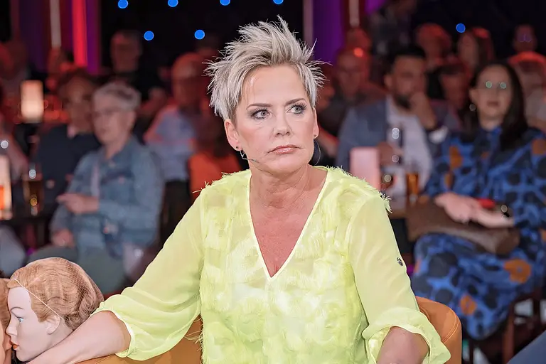 Inka Bause, hier in einer gelben Bluse und einer grünen Hose zu sehen, sieht sehr ernst aus. Sie trauert um ein Teammitglied.