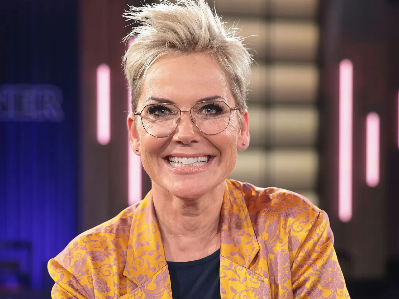 Inka Bause trägt einen bunten Blazer und eine Brille. Sie strahlt in die Kamera.