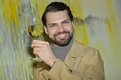 Jimi Blue Ochsenknecht trägt einen beigen Trenchcoat und hält grinsend ein Glas Weißwein hoch.