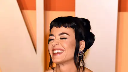 Katy Perry lächelt schräg an der Kamera vorbei und hat die Hände in die Hüfte gestemmt. Sie trägt ein goldenes Abendkleid und hat einen kurzen Pony
