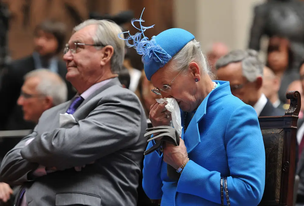 Königin Margrethe im blauen Kostüm mit passendem Hut sitzt auf einem Stuhl und putzt sich die Nase. Königin Margrethe im blauen Kostüm mit passendem Hut sitzt auf einem Stuhl und putzt sich die Nase.