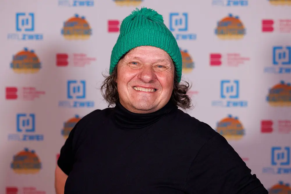Manni Ludolf trägt eine grüne Mütze und ein schwarzes Oberteil mit Rollkragen. Er grinst in die Kamera. Manni Ludolf trägt eine grüne Mütze und ein schwarzes Oberteil mit Rollkragen. Er grinst in die Kamera.