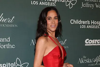 Meghan in einem roten Abendkleid mit weitem Ausschnitt und Trägern. Sie schaut zur Seite und sieht halb glücklich, halb erschrocken aus. 