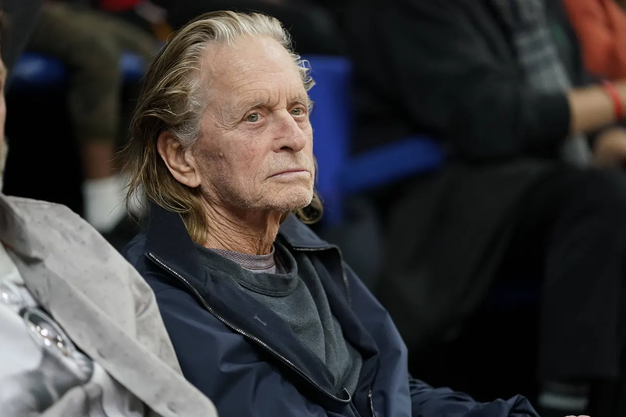 Michael Douglas sitzt mit ernster Miene bei einem Basketballspiel. Seine langen Haare hat er sich aus dem Gesicht gekämmt. - Foto: IMAGO / Panoramic by PsnewZ