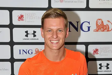 Mick Schumacher bei einem Event. Er trägt ein Oranges Muskelshirt von Under Armour. Er schaut direkt in die Kamera und hat ein strahlendes Lächeln auf dem Gesicht. 