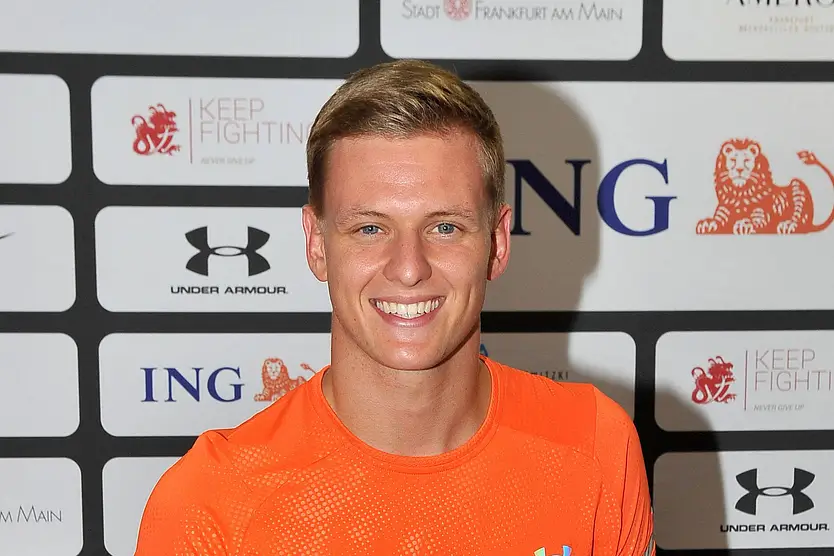Mick Schumacher bei einem Event. Er trägt ein Oranges Muskelshirt von Under Armour. Er schaut direkt in die Kamera und hat ein strahlendes Lächeln auf dem Gesicht. 