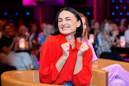 Mimi Fiedler trägt eine rote Bluse und hält grinsend ihre Arme vor die Brust. 