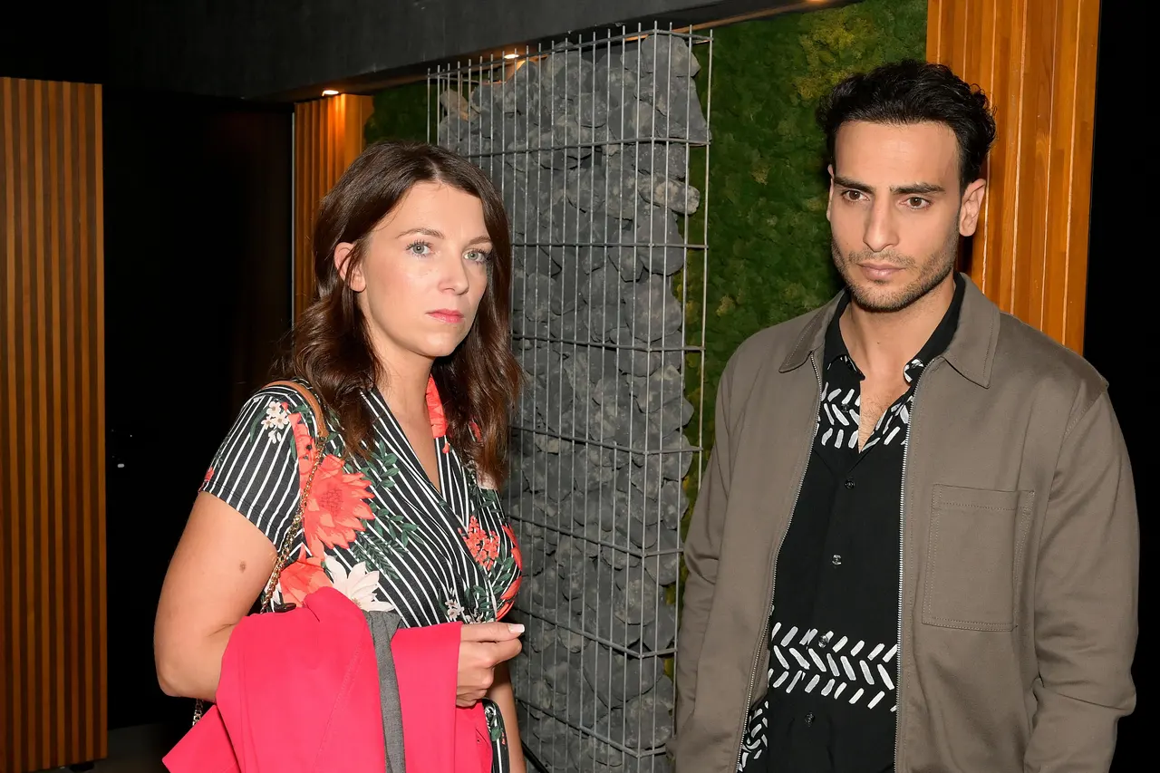 Nihat in grauer Jacke und Lilly in einem Kleid mit Blumenmuster schauen bedrückt in die Ferne.