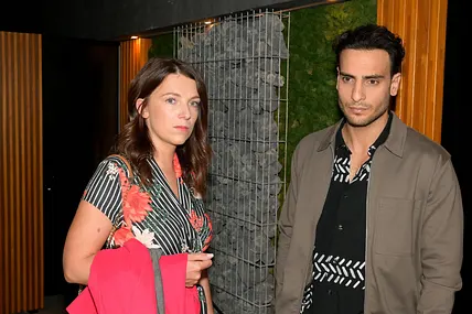 Nihat in grauer Jacke und Lilly in einem Kleid mit Blumenmuster schauen bedrückt in die Ferne.