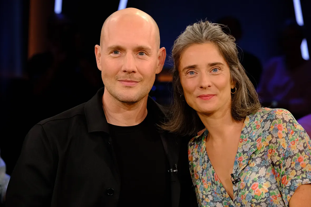 Oli.P, mit Glatze und in Schwarz gekleidet, neben seiner Frau Pauline, die ein geblümtes Kleid trägt.