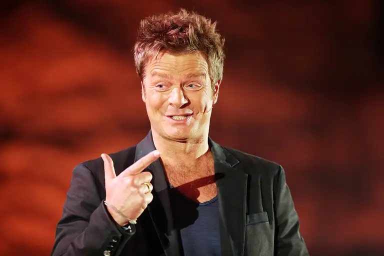 Oliver Geissen lächelt und hält zwei Finger in die Höhe.