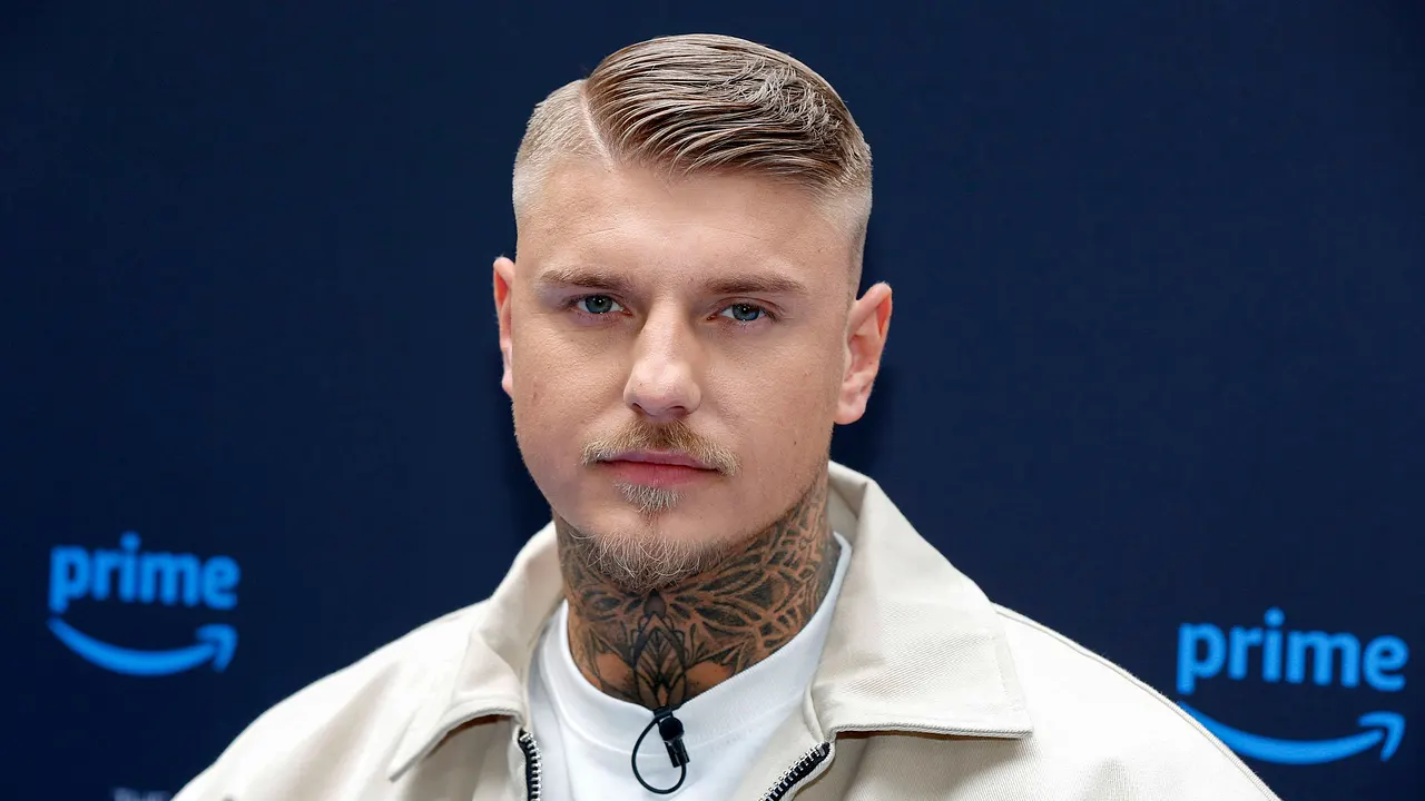 Paco Herb in weißem T-Shirt mit beiger Jacke auf dem Teppich von der Premiere von "The 50", er hat einen durchdringenden ernsten Blick