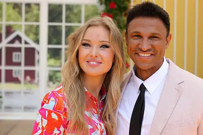 Patrice Aminati und ihr Mann Daniel Aminati lächeln in die Kamera. Sie trägt ein buntes Kleid, er einen rosafarbenen Anzug. 