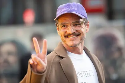 Pedro Pascal trägt eine Brille und eine Kappe. Er lächelt und macht ein Peace-Zeichen mit den Fingern.