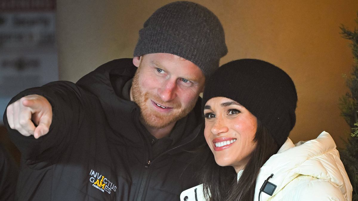 Prinz Harry zeigt mit dem rechten Zeigefinger vor sich, Meghan blickt ebenfalls lächelnd nach vorne.