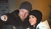 Prinz Harry zeigt mit dem rechten Zeigefinger vor sich, Meghan blickt ebenfalls lächelnd nach vorne. - Foto: IMAGO / Cover-Images