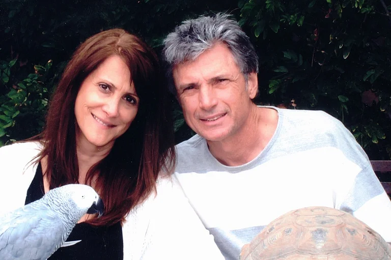 Rpbin Kaye und Thomas Deluca sitzen mit einem Papagei und einer Schildkröte im Garten und lächeln in die Kamera.